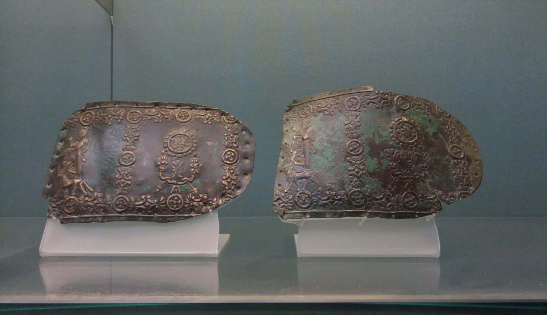 Fragments of an Urartian bronze belt on museum mounts, embossed with banded borders, rosette discs, and small figural scenes of deities standing on a bull and a lion.